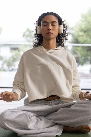 Non-binary person meditating with headphones in cream hoodie and gray sweatpants