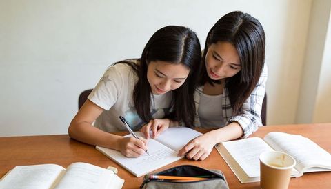 Asian female students studying together writing notes and collaborating at wooden desk