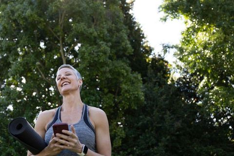 Mature woman enjoying outdoor yoga with smartphone and rolled mat under leafy canopy