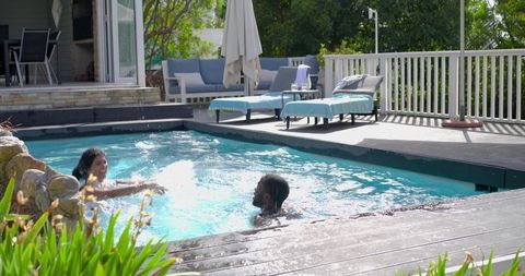 Diverse couple splashing in backyard pool with wood deck, lounge chairs and stone feature