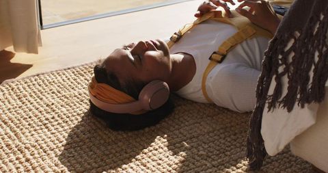Sunlit asian woman wearing headphones lying on jute rug cozy living room wellness calm