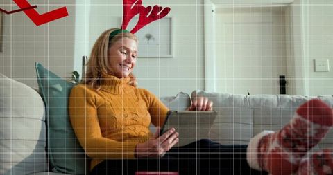 Mature woman relaxing on couch wearing reindeer antlers and using tablet in festive mood