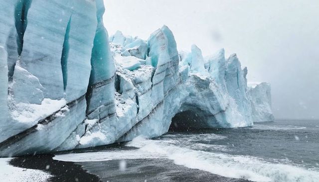 Towering blue glacier sea arch and black sand shoreline in falling snow