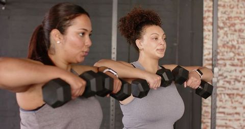 Diverse women doing upright rows with hex dumbbells in industrial brick gym studio