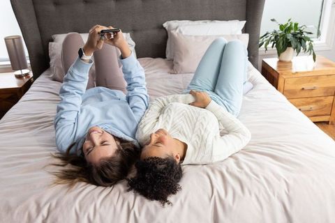 Multicultural friends relaxing and scrolling smartphone together on cozy bedroom bed