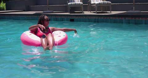 African american woman relaxing on pink donut float in turquoise pool wearing sunglasses