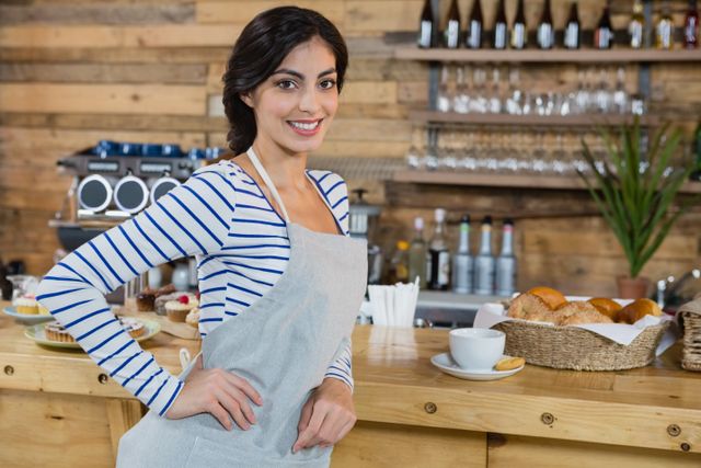 Smiling waitress standing confidently at counter in cafeteria. She wears an apron and stripes shirt, behind her are shelves with glasses and pastries. Ideal for hospitality, service industry, and small business themes. Great for websites, promotional materials, or articles related to customer service, cafes, and small businesses.