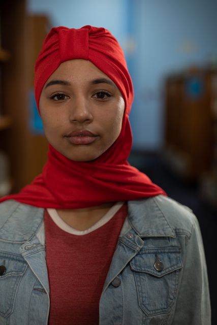Portrait of an Asian female student wearing a red hijab and jeans jacket studying in a library standing, looking straight to camera.