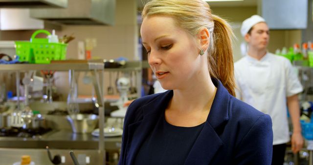 A professional woman is seen carefully inspecting an industrial kitchen, indicative of restaurant or commercial kitchen management. In the background, a chef in uniform appears focused on his tasks. This can be used to illustrate themes related to restaurant management, food safety inspections, team coordination, or culinary training. It is ideal for articles or advertisements showcasing the behind-the-scenes operations of the foodservice industry.