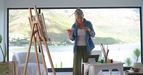 Mature artist painting at easel in coastal studio using tablet for reference