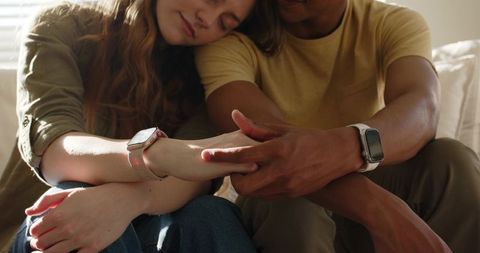 Couple wearing smartwatches sharing intimate daylight moment on sofa, connected wellness lifestyle
