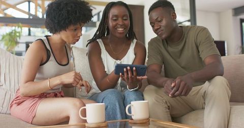 Black friends laughing and sharing phone on couch in bright living room