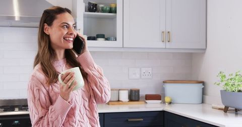 Smiling woman talking on smartphone with coffee mug in modern home kitchen