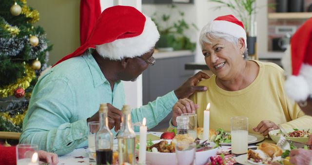 Happy diverse senior couple in santa hats talking at christmas dinner table at home. retirement lifestyle, christmas festivities, celebrating at home with friends.