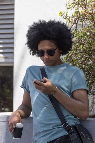 African american man with afro leaning on wall checking smartphone, holding coffee