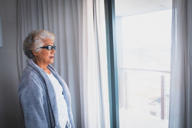 Senior woman standing by window, wearing glasses and robe, looking outside. Ideal for topics on senior lifestyle, retirement, quarantine, isolation, and elderly care. Can be used in articles, blogs, and advertisements focusing on senior living, mental health, and home life.