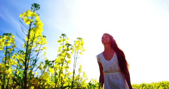 A woman is enjoying a sunny day in a rapeseed field, surrounded by yellow blossoms. She is smiling widely, dressed in a white dress under a clear blue sky. This image can be used in contexts promoting wellness, outdoor activities, lifestyle blogs, and ads focused on nature, freedom, and happiness.