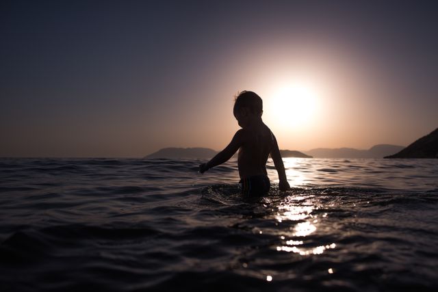 A young boy, silhouetted by the setting sun, plays in the ocean water. The picturesque sunset creates a peaceful backdrop, highlighting the ripples in the water and the distant horizon. This depiction of childhood joy and exploration is perfect for themes related to vacations, summer activities, tranquil outdoor settings, and the beauty of seaside adventures.
