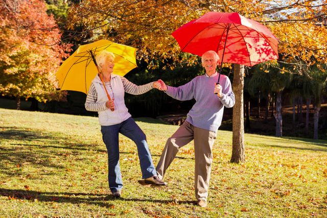 Senior couple enjoying a playful dance with colorful umbrellas in an autumn park. Perfect for themes related to active aging, retirement, happiness, and outdoor activities. Ideal for use in advertisements, brochures, and articles promoting senior wellness and active lifestyles.