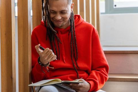 African american man journaling on wooden stairs wearing red hoodie and dreadlocks