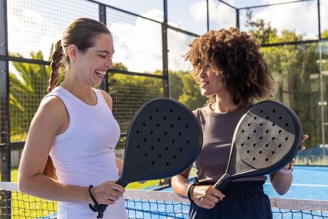 Smiling diverse women sharing padel paddles on sunlit outdoor court, friendly match moment