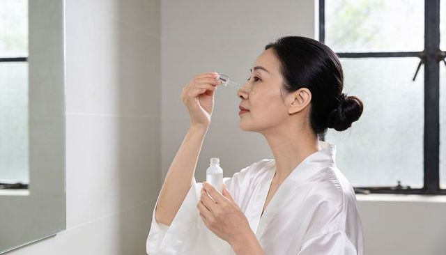 Asian woman applying hydrating facial serum with glass dropper at bathroom vanity