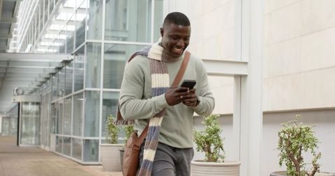 African american student walking on modern campus checking phone with scarf and satchel