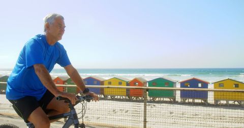 Senior Man Biking by Beachfront with Colorful Huts in Background