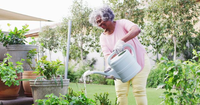 Senior woman wearing casual clothes watering plants with a watering can in a garden. She is surrounded by potted plants and greenery, enjoying an outdoor activity. Use for articles on active aging, gardening tips, retirement lifestyle, or health benefits of outdoor activities.