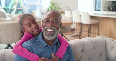 Grandfather and granddaughter sharing joyful hug on tufted sofa in bright family living room