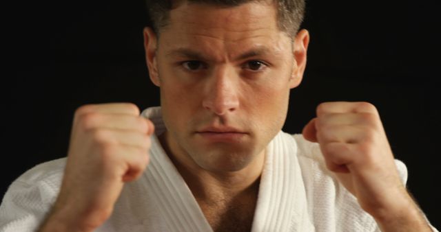 Martial artist dressed in white uniform with intense look on face, holding fists up. Useful for illustrating concepts of determination, self-discipline, self-defense principles, or martial arts training programs.