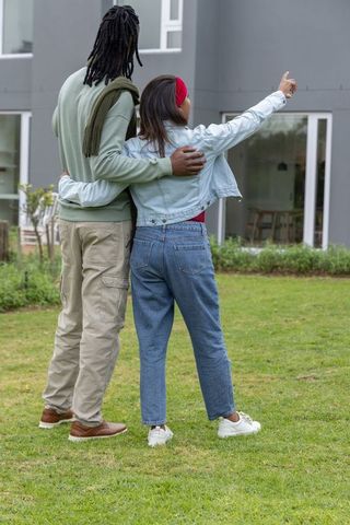 Diverse couple choosing new home outside modern house, woman pointing, embracing