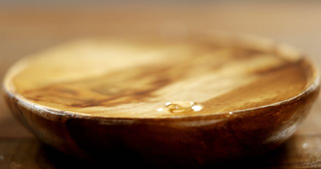 Closeup showing a wooden bowl containing olive oil, focusing on the smooth texture and rich brown tones of the wood, enhanced by the oil's reflection. Ideal for use in articles, blogs, and websites about cooking, natural materials, organic products, and rustic home decor concepts.