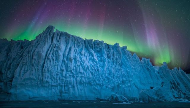 Rising blue glacial cliff under dancing aurora borealis and starry polar night sky