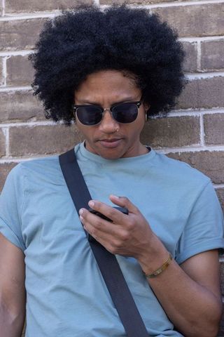 African american man leaning on brick wall checking smartphone wearing casual sunglasses
