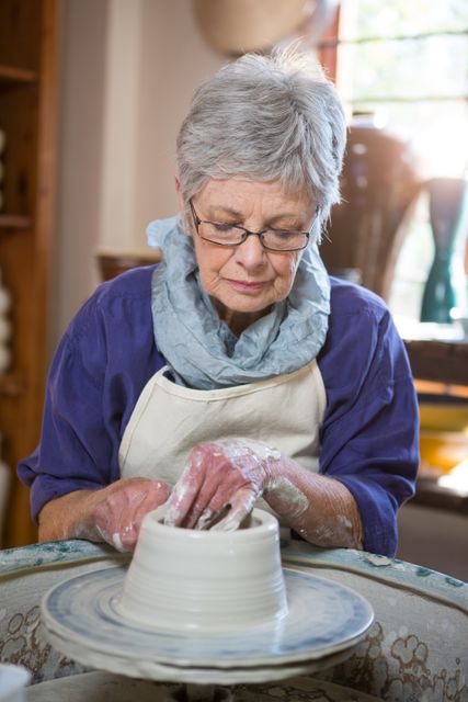 Senior woman shaping clay on pottery wheel in a workshop. Ideal for blogs, articles, or advertisements focused on arts and crafts, senior hobbies, creative activities, or pottery classes. Highlights craftsmanship, creativity, and traditional art forms.
