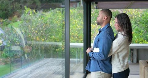 Couple embracing by glass wall overlooking garden, african american man holding letter