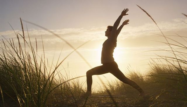 Practicing warrior ii yoga on coastal dune at golden hour silhouette stretching