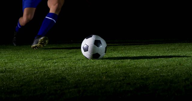 A soccer player in blue gear is about to kick a ball on a lush green pitch at night, with copy space. Capturing the dynamic action of the sport, the image conveys the intensity and focus of a soccer match under the lights.