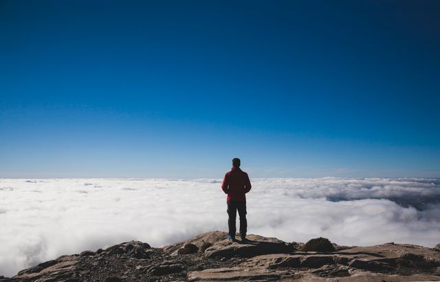 Solitary hiker standing at mountain peak overlooking expansive cloudscape. Perfect for themes around adventure, travel, outdoor activities, and the serenity of nature. Ideal for blogs, inspirational posters, travel websites, and promotional materials emphasizing exploration and peacefulness.