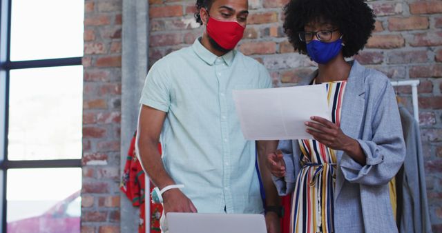 Diverse male and female fashion designers wearing face masks in discussion at work looking at paper. independent creative design business during coronavirus covid 19 pandemic.