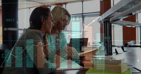 Two businesswomen collaborating over documents and laptop with financial charts overlay in library