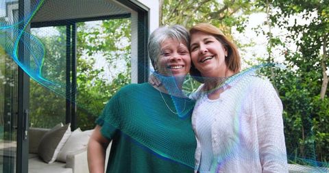 Mature women smiling on garden patio leaning by glass door, embracing, lifestyle portrait