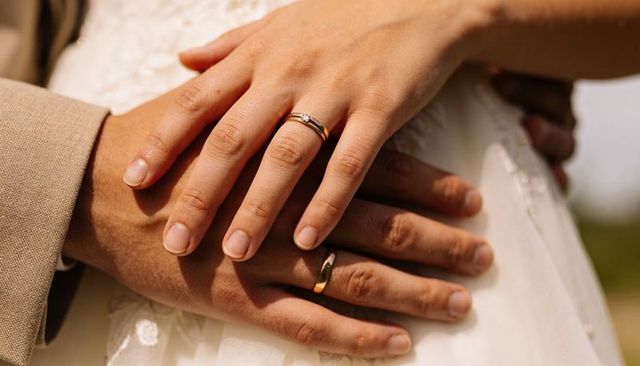 Newlywed hands embracing with wedding rings and lace dress detail in sunlit outdoor moment