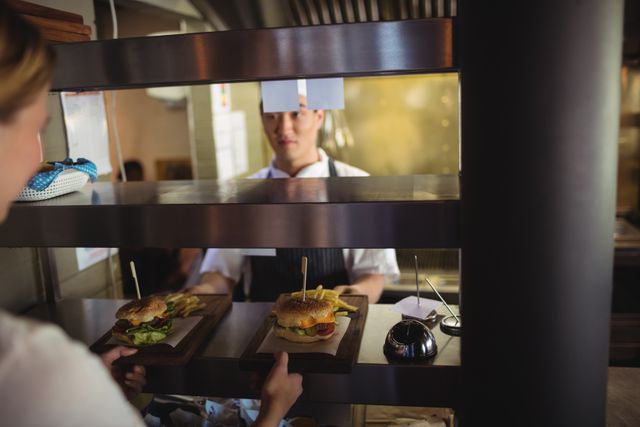 Chef handing over a tray with a burger and french fries to a waitress in a busy commercial kitchen. Ideal for use in articles or advertisements related to restaurant operations, food service, culinary arts, and the hospitality industry. Can also be used for content focusing on teamwork and professional kitchen environments.