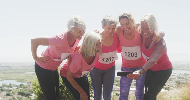 Group of senior women taking a selfie after participating in a breast cancer awareness race, all wearing pink shirts and race numbers. Image is suitable for campaigns promoting health, awareness, and support for cancer fundraising events, celebrating female friendships and community spirit.