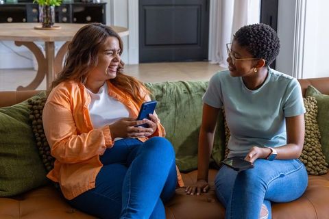 African american friends laughing and sharing smartphones while sitting on cozy leather couch