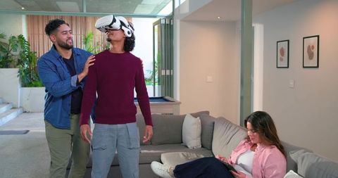 Friends Guiding Man Testing Virtual Reality Headset in Modern Living Room, Woman Using Tablet