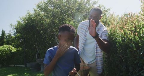 African american father gardening with teenage son holding basket in sunny backyard