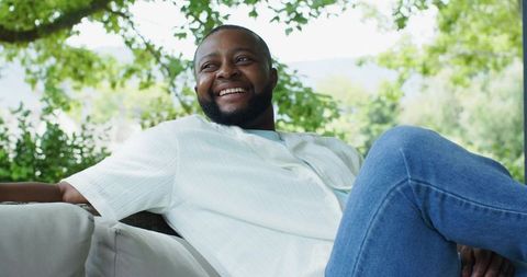 Smiling man relaxing on outdoor sofa under trees wearing short-sleeve shirt and jeans